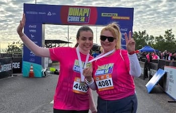 Dos mujeres con camisetas rosas sonríen, una muestra su medalla y hace un gesto de paz. Ambiente festivo con arco de meta detrás.