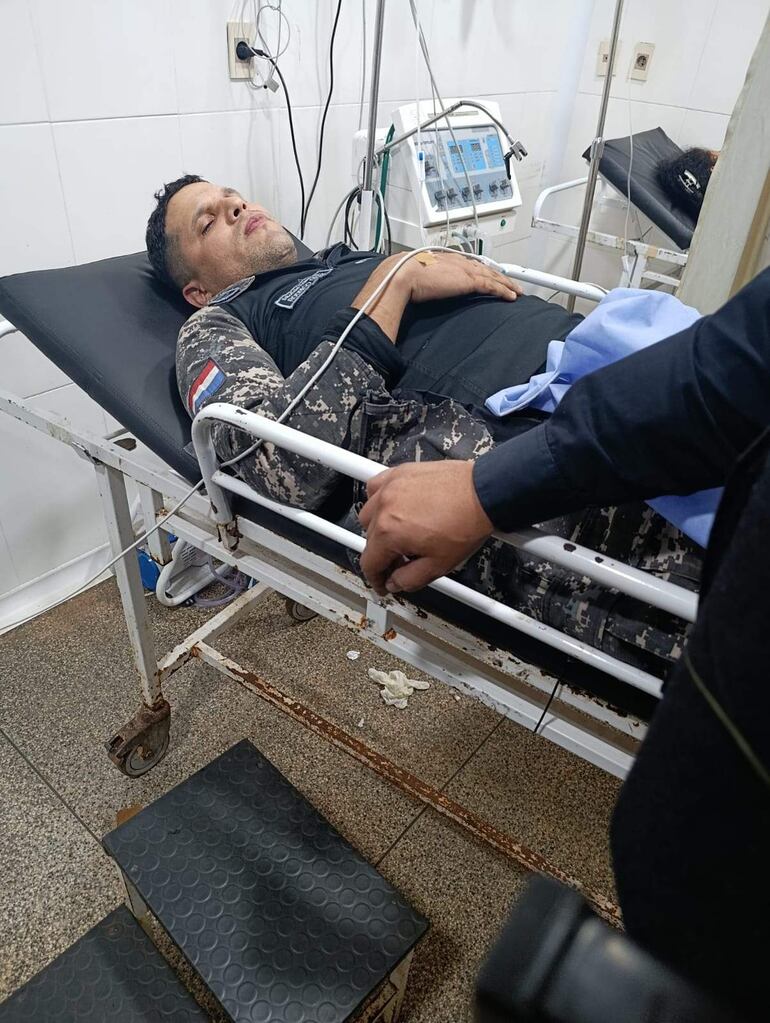 Hombre en camilla con uniforme de camuflaje y bandera paraguaya, con expresión de malestar en un entorno hospitalario.
