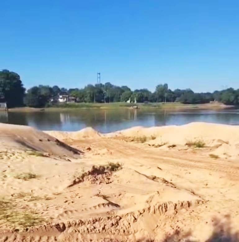 Paisaje ribereño despejado con cuerpo de agua y arena en la orilla, rodeado de vegetación y cielo azul.
