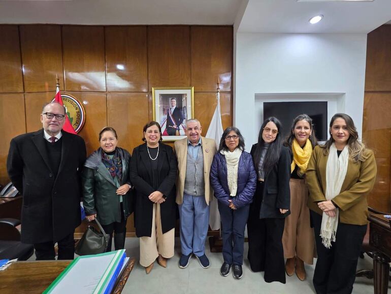 Grupo de siete personas sonrientes en ambiente oficial, con banderas y retratos en el fondo, vestidos de manera formal.