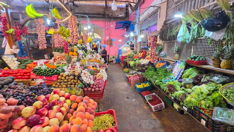 Comerciante en un mercado iluminado atendiendo a cliente, rodeado de coloridas frutas y verduras frescas.