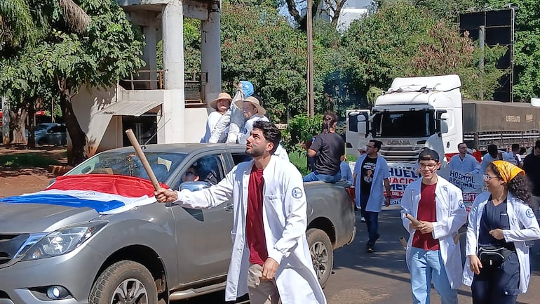 Médicos en batas blancas sosteniendo un palo y protestando, con bandera paraguaya visible en una camioneta detrás.