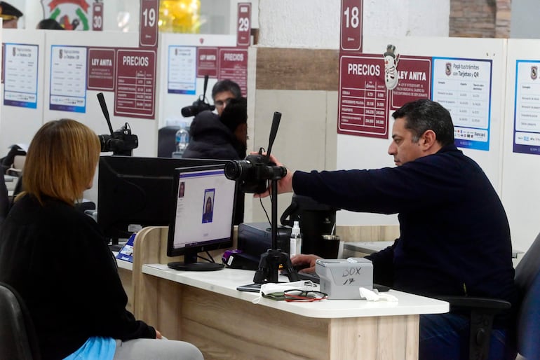 Mujer con suéter negro sentada y hombre de pie tomando una fotografía, en entorno de oficina con objetos de trámite.