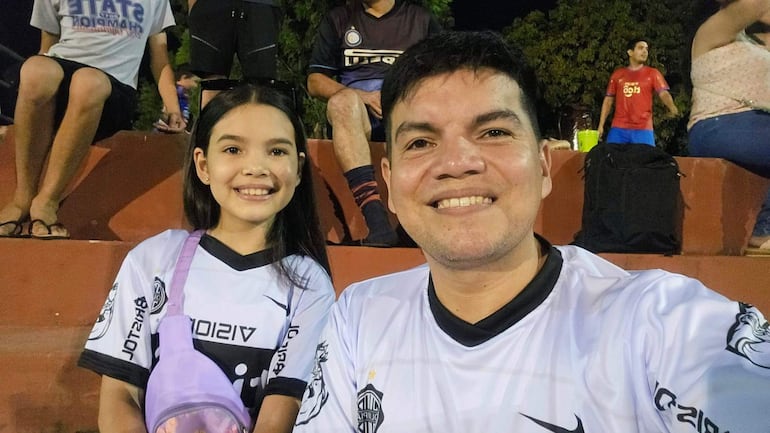 Hombre sonriendo en primer plano, niña con mochila lilac y camiseta de fútbol, ambos en un ambiente de grada deportiva.