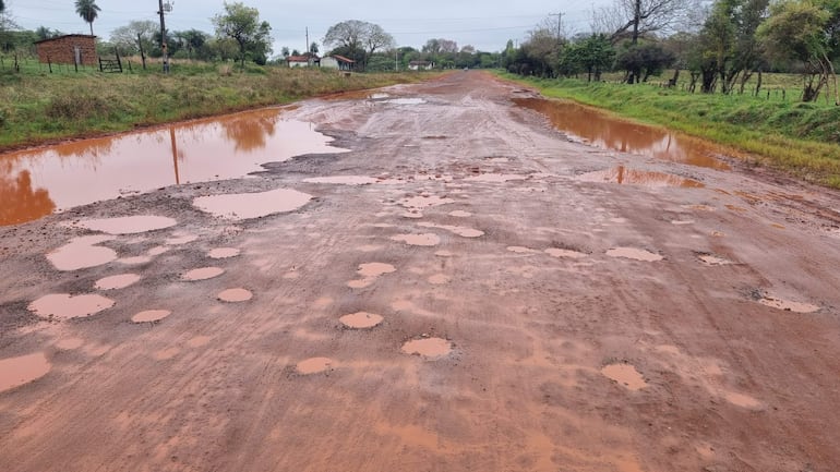Carretera rural en mal estado con barro y charcos, rodeada de vegetación y pequeñas construcciones.