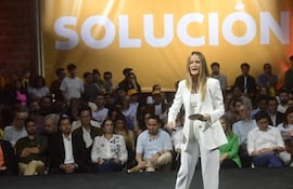 Soledad Núñez en traje blanco, hablando al público con expresión enérgica, fondo amarillo con la palabra 'SOLUCIÓN'.