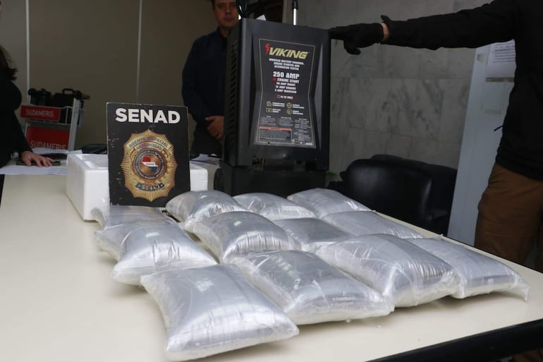 Emblema de la Senad en primer plano, mujer atenta y hombre con batidor de batería en entorno serio y profesional.