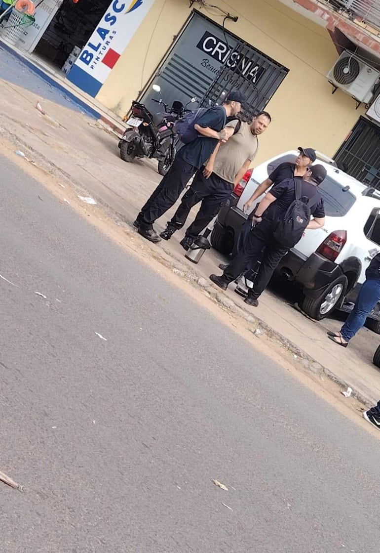 Cinco hombres en la acera: uno con botella de acero y otros conversando, en un entorno urbano con comercios al fondo.