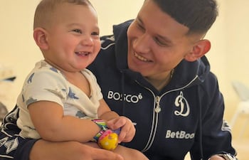 Juan Cáceres sonríe mientras sostiene a un bebé en un sofá, ambos luciendo alegres en un ambiente acogedor.