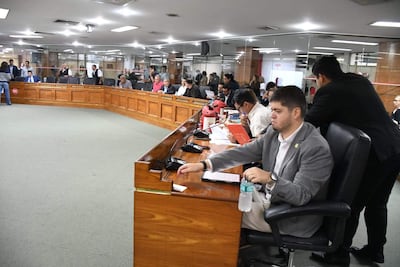 Juan Carlos Ozorio Admen sentado con documentos, mientras Miguel Sosa se encuentra de pie, en un ambiente formal con otras personas observando.