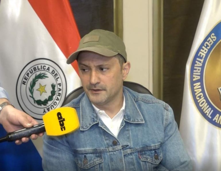 Jalil Rachid sentado en silla, con chaqueta de mezclilla y gorra beige. Una mano sostiene un micrófono de ABC en un entorno con banderas de Paraguay.