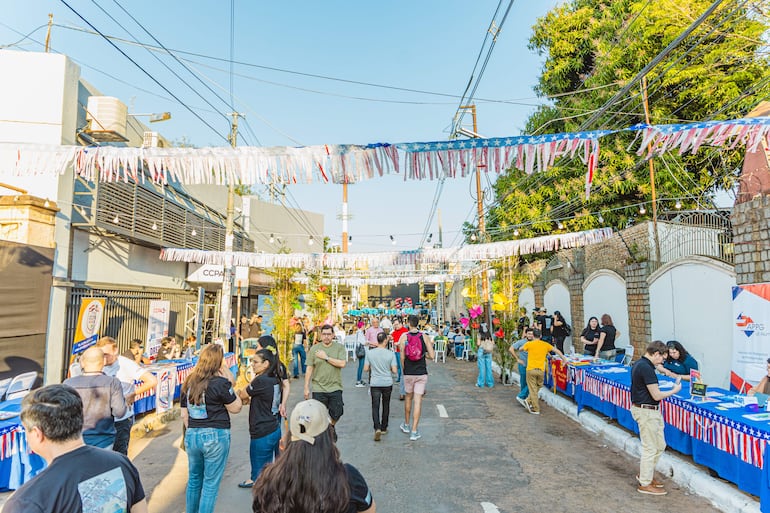 Personas socializando en una calle con banderines rojo, azul y blanco, en un ambiente festivo al aire libre.