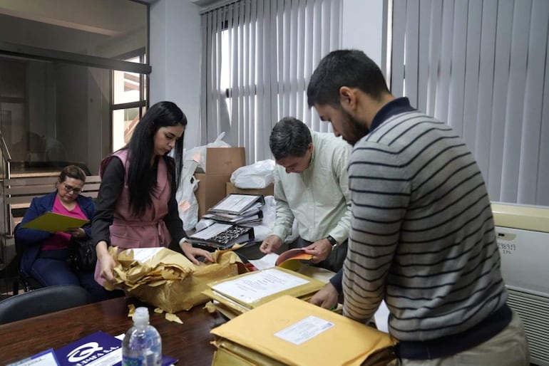 Dos hombres y dos mujeres en oficina revisan documentos. Una mujer abre un paquete, mientras otro hombre examina papeles en la mesa.