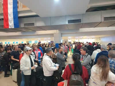 Multitud de aficionados con camisetas de Paraguay en fila en un aeropuerto, algunos sonrientes y otros conversando.