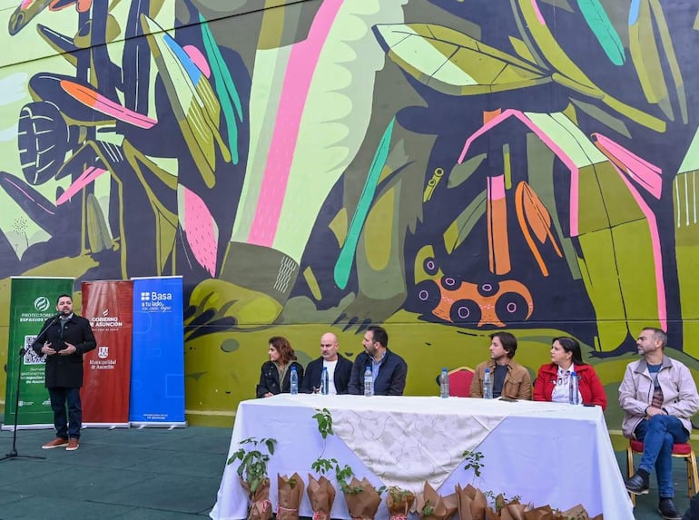 Luis Bello habla en un micrófono, rodeado de cinco personas atentas, en un evento al aire libre decorado con un mural colorido.