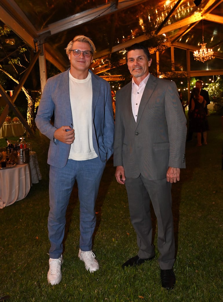 Hombres en trajes, uno claro con zapatos deportivos y otro gris con zapatos oscuros, sonríen en césped decorado con luces.