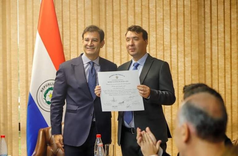 Hombre sonriente en traje oscuro junto a otro hombre con documento, aplaudido en ceremonia con bandera paraguaya al fondo.