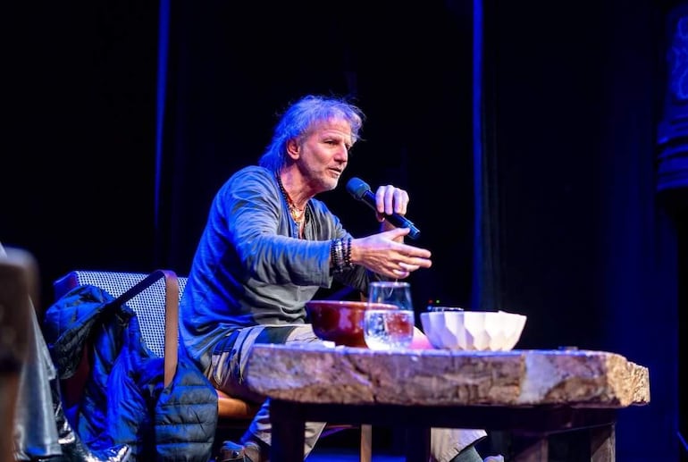 Facundo Arana sentado en un escenario, vestido casual, sosteniendo un micrófono y gesticulando con expresión tranquila.