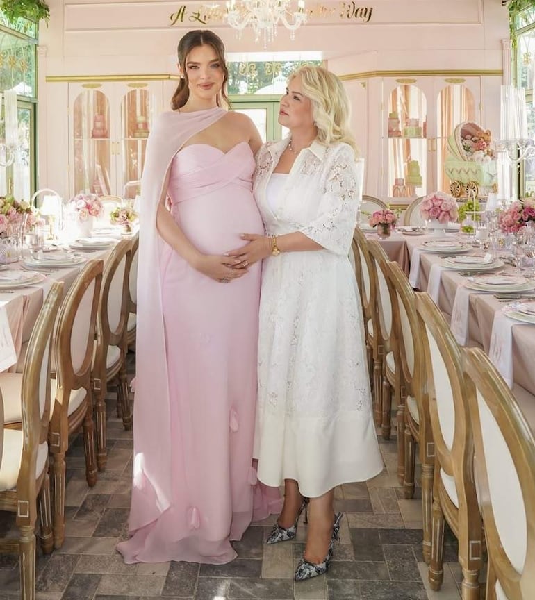Nadia Ferreira en vestido rosa, sonriente y embarazada, conversa con mujer vestida de blanco en salón decorado con flores.
