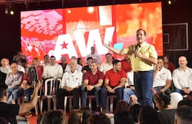 Arnaldo Samaniego en camisa amarilla, de pie con micrófono, rodeado de personas vestidas en blanco y rojo, escuchando atentos.