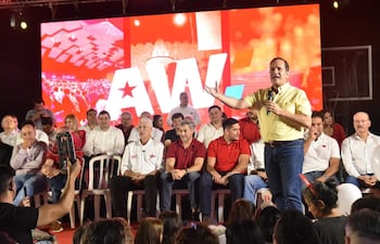 Arnaldo Samaniego en camisa amarilla, de pie con micrófono, rodeado de personas vestidas en blanco y rojo, escuchando atentos.