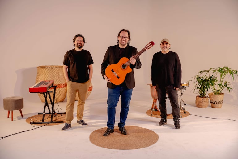 Músicos sonrientes en estudio: uno en blazer negro sostiene la guitarra, otro usa suéter negro y gorra, en ambiente decorado con plantas.