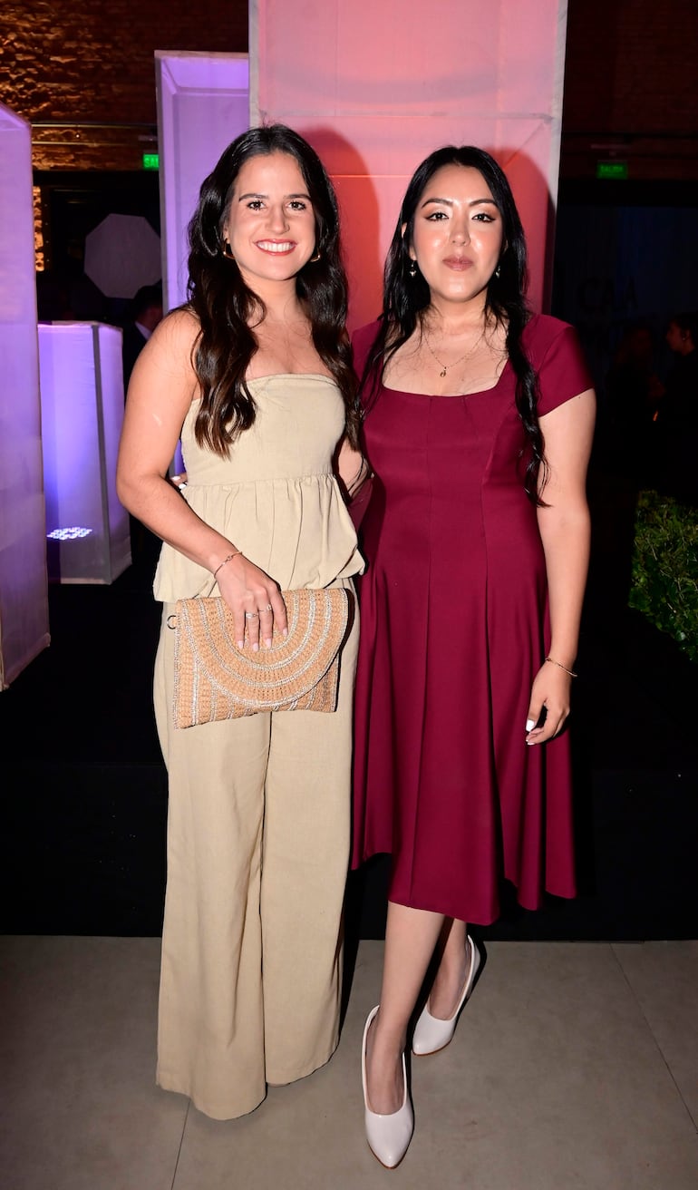 Dos mujeres, una en vestido burdeos y otra en atuendo claro, sonríen en una elegante gala con decoración morada.