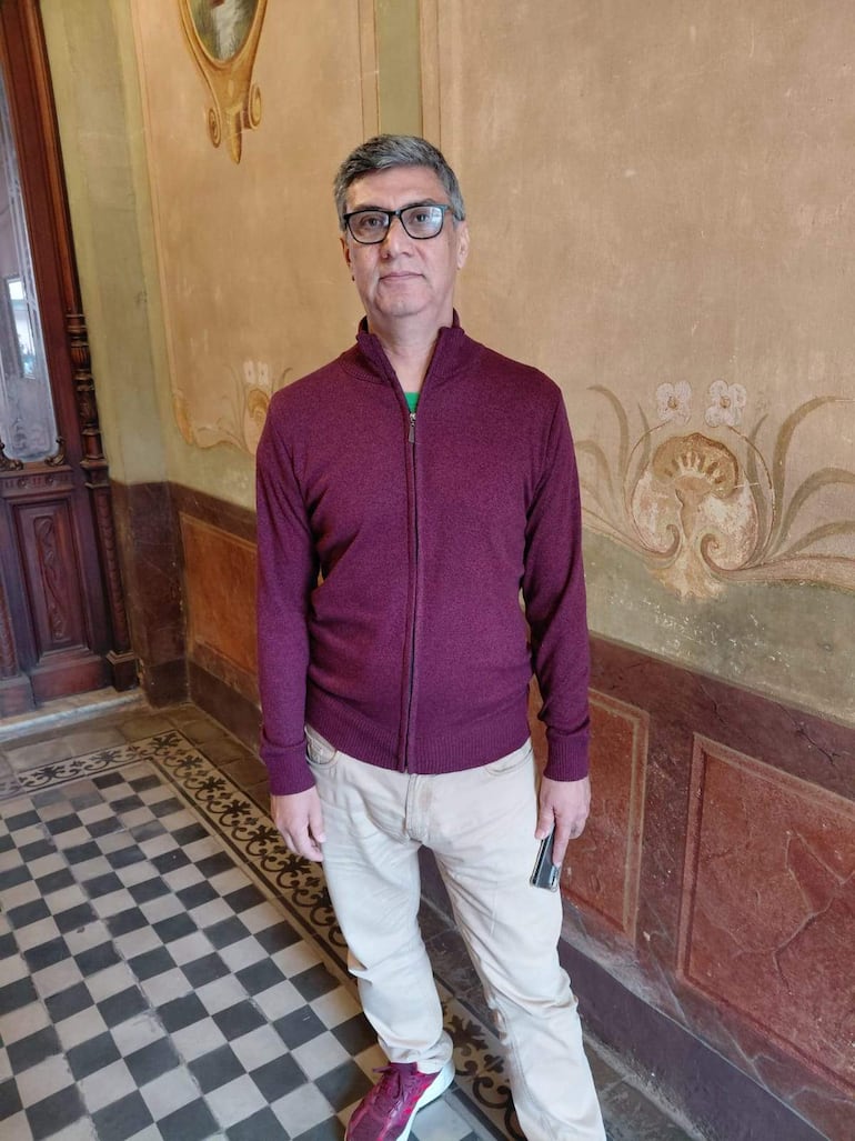 Hombre de pie con suéter burdeos y pantalones claros, en un elegante ambiente interior, observando a la cámara.