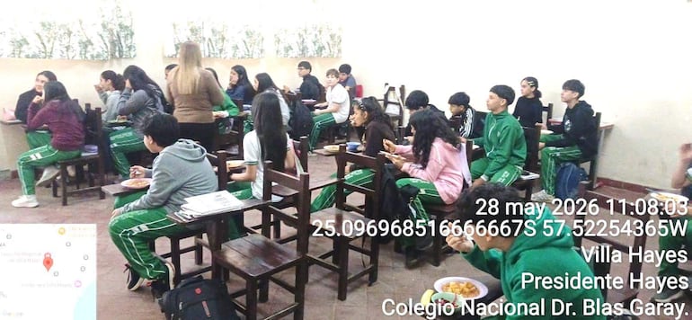 Estudiantes con camisetas blancas y pantalones verdes conversan y comen en un aula, con mesas de madera alrededor.