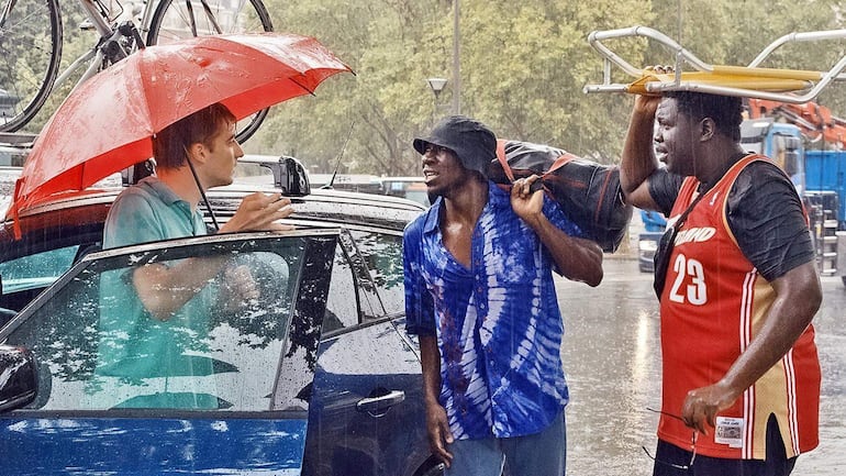Hombre con paraguas rojo y camiseta número 23 conversa con dos hombres empapados y con mochilas bajo la lluvia.
