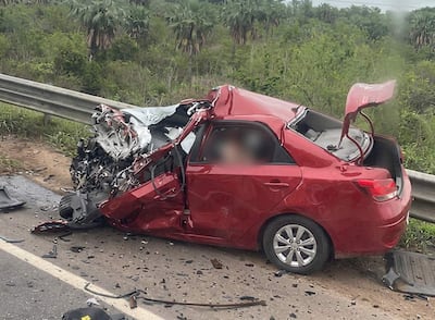 Automóvil rojo gravemente dañado tras accidente en ruta, con partes esparcidas y puerta abierta, en un entorno natural.