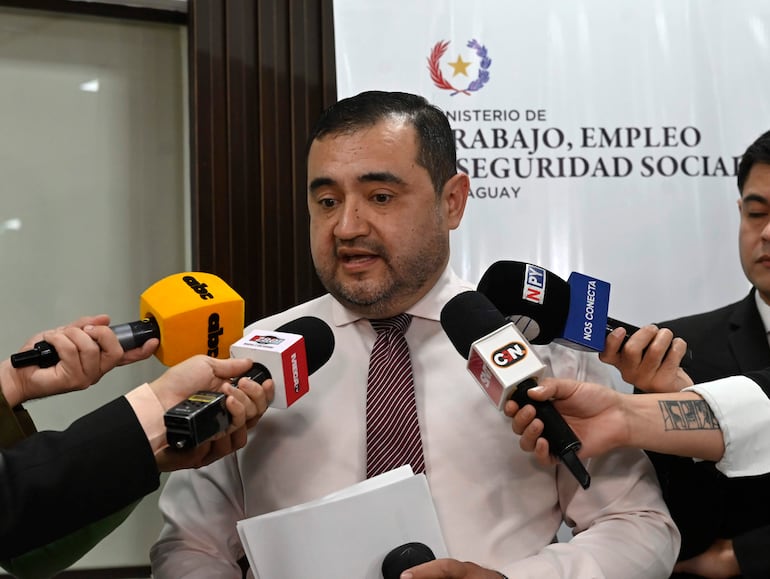 Hombre de cabello oscuro, camisa clara y corbata de rayas, habla ante micrófonos sostenidos por varias manos de periodistas en un entorno formal.