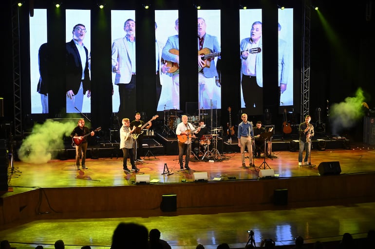 Músicos en el escenario con luces verdes; el vocalista principal con guitarra y camisa blanca, acompañado de otros músicos.