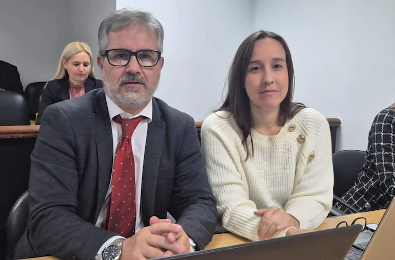 Carlos Raúl Arce Letelier con traje oscuro y corbata roja, junto a mujer con suéter blanco, en una sala de reuniones.