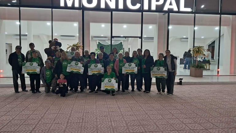 Un grupo de adultos mayores en camisetas verdes sostiene carteles en la entrada de un edificio municipal, expresando diversas emociones.