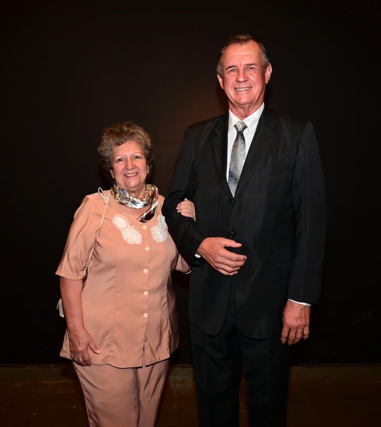 Luis Christ Jacobs sonríe en traje oscuro, mientras una mujer a su lado viste blusa clara y pañuelo, ambos en evento formal.