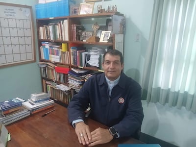 Monseñor Gabriel Escobar sentado en una oficina, con camisa blanca y chaqueta oscura, revisa un calendario en su escritorio.
