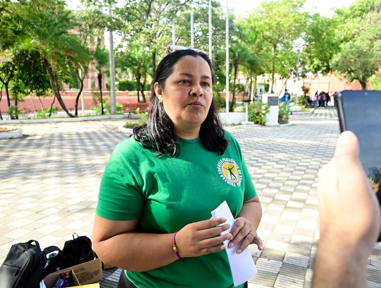Mujer con cabello largo y lacio, camiseta verde, sostiene un papel y conversa en un parque con árboles alrededor.