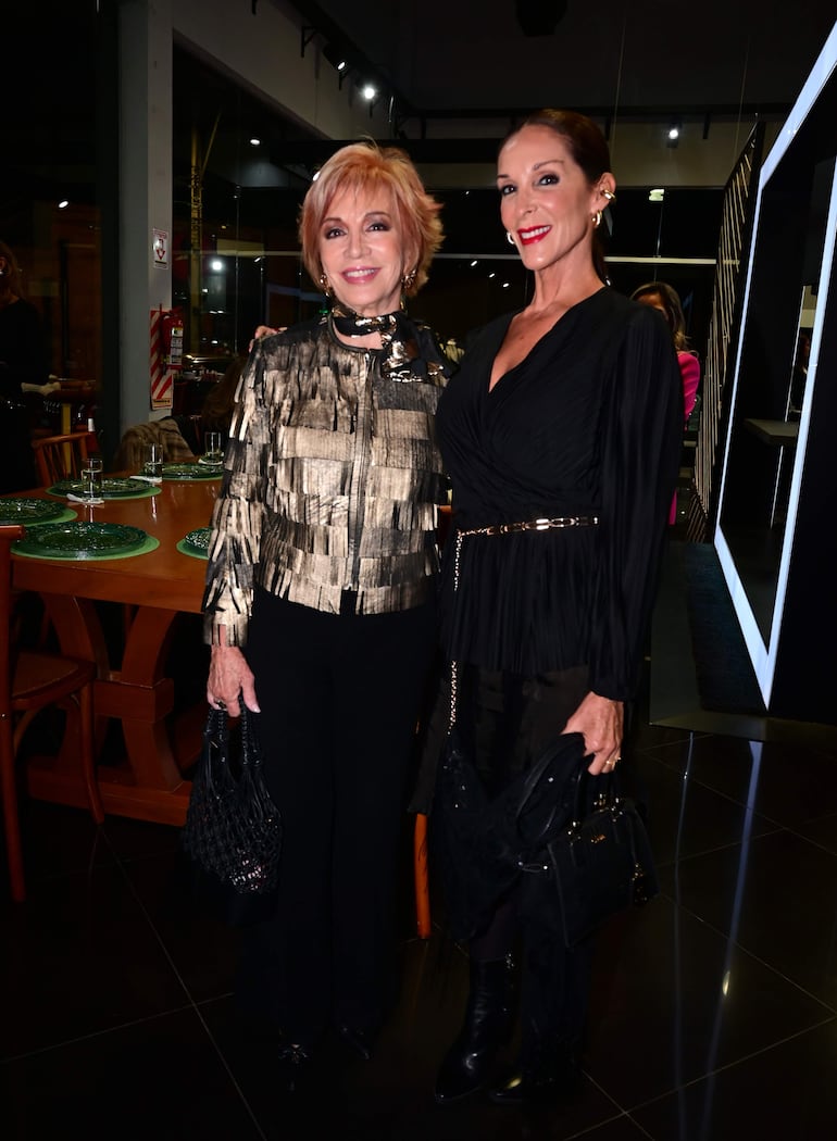 Dos mujeres sonrientes, una con abrigo dorado y negro y otra con vestido negro, posan en elegante ambiente festivo con mesas decoradas.