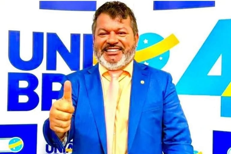 Aparecido Carlos Bernardo sonríe con traje azul, camisa y corbata amarillas, levantando el pulgar en ambiente con el texto 'Unidos Brasil'.
