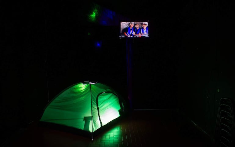 Tienda de campaña verde iluminada, televisor mostrando personas no identificadas, sillas apiladas en un ambiente oscuro.