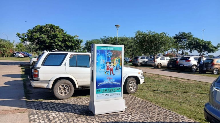 Estacionamiento abarrotado de vehículos junto a un gran cartel publicitario de 'Disney On Ice' en un ambiente verde.