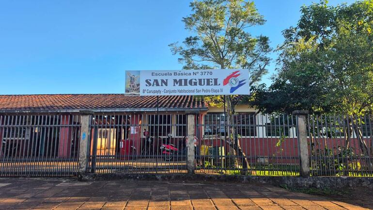 Fachada de la Escuela Básica N° 3770 'San Miguel' con cartel y rejas, rodeada de árboles en un día despejado.