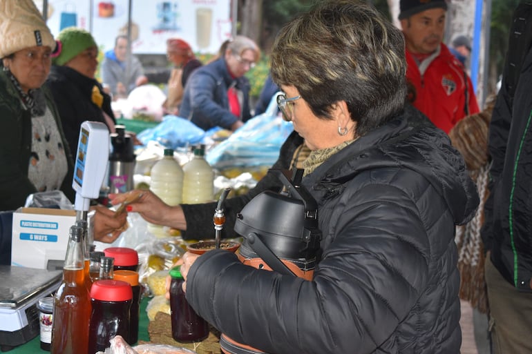 Mujer de cabello corto y anteojos sostiene una jarra, realizando una transacción con otra mujer en el mercado al aire libre.