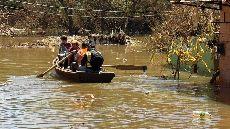 Niño y hombre con sombrero reman en canoa, mientras un hombre con chaqueta naranja ayuda. Agua y desechos alrededor.