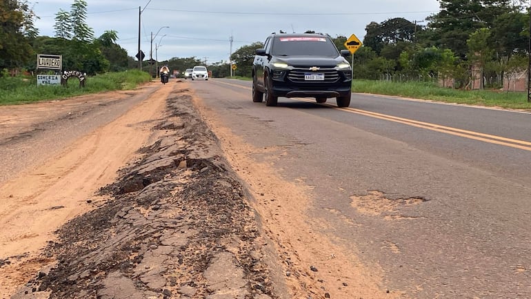 Ruta PY05 con asfalto agrietado y arena, mientras un auto Chevrolet transita. Cartel de 'Lavadero' visible a la izquierda.