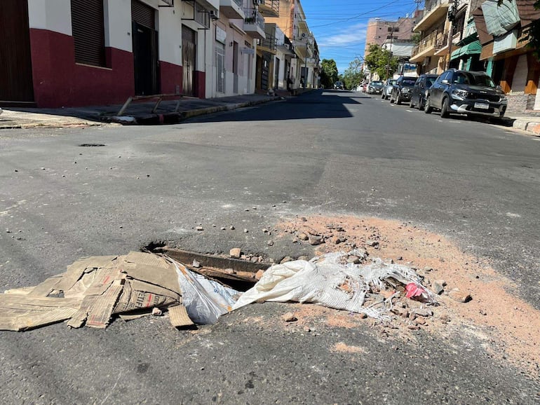 Calle vacía con gran bache rodeado de escombros, edificios de varios pisos al fondo bajo luz diurna.