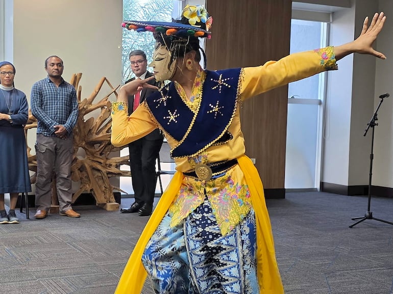 Hombre en traje colorido bailando en una celebración cultural, con tres observadores atentos en un entorno formal de madera.