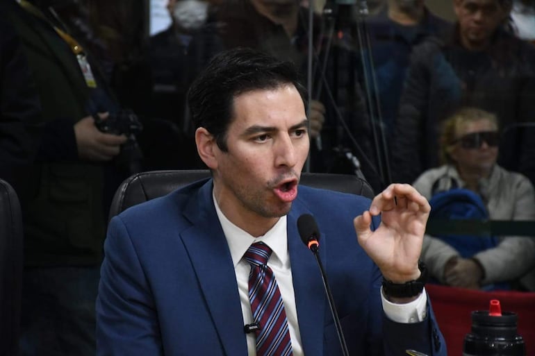 Álvaro Grau en traje azul y corbata de rayas, gesticulando frente a un micrófono y una multitud de asistentes detrás.
