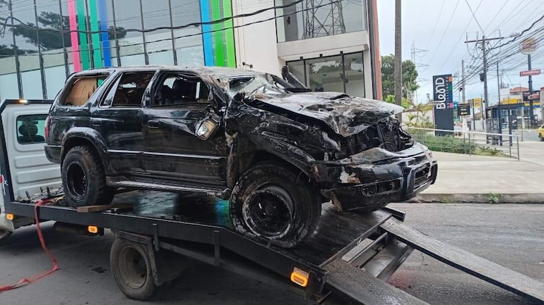 Camioneta negra gravemente dañada en una plataforma de camión, en un entorno urbano con un edificio moderno al fondo.